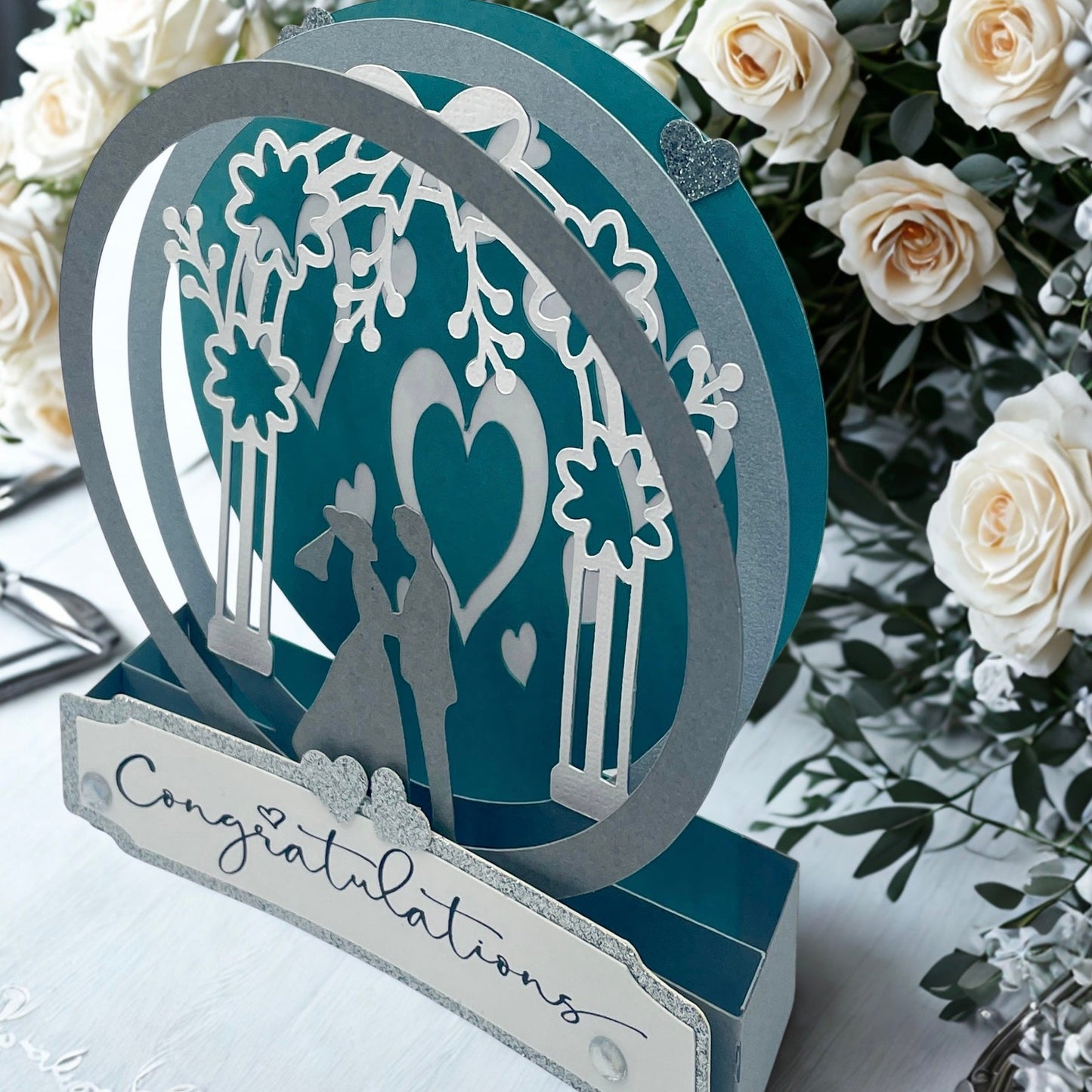Decorative wedding card with cut-out design of a couple under an arch, surrounded by white roses.