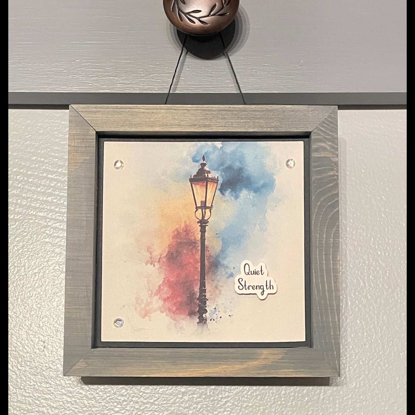 Framed artwork with a lamp post and 'Quiet Strength' text on a textured wall.