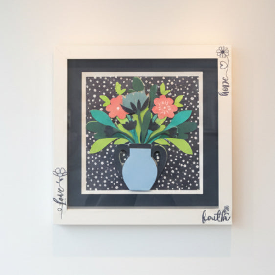 Framed artwork of a floral arrangement with a blue vase on a polka dot background.
