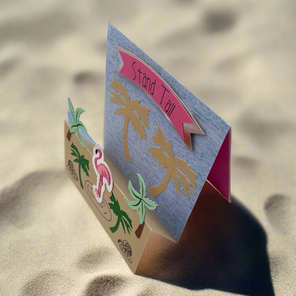 A greeting card with a stand-up design featuring a flamingo and palm trees on a background that suggests a tropical setting. The front of the card reads 'STAND TALL' with a pink banner and the inside message is 'Because you are Flamazing!'.
