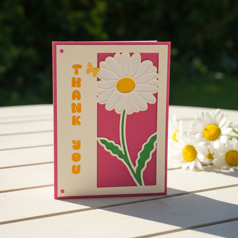 Card with a daisy design and 'Thank You' text on a wooden surface with flowers.