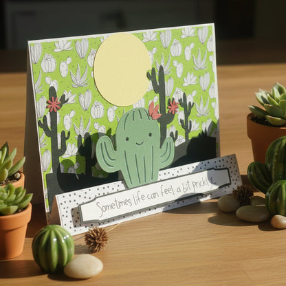 Card with cactus design on a wooden surface with potted plants and stones.