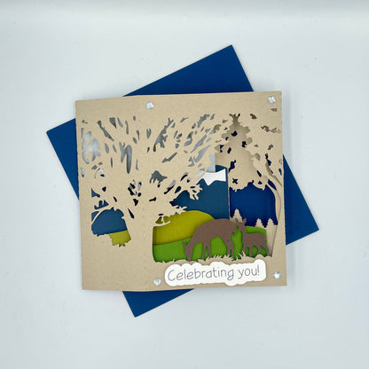 Card with cut-out design of a bear and trees, featuring the text 'Celebrating you!' on a blue envelope.