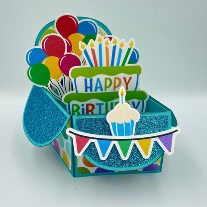 Colorful birthday-themed gift box with balloons, cake, and cupcake decorations on a light gray background.