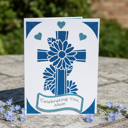 Greeting card with blue cross design and 'Celebrating You, Mom' text on a stone surface with flowers.