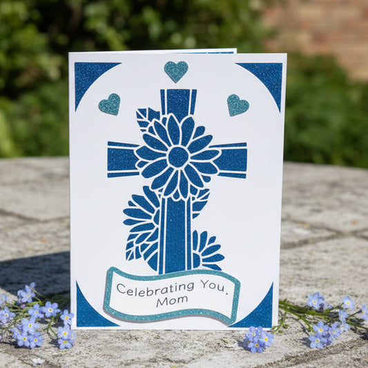 Greeting card with blue cross design and 'Celebrating You, Mom' text on a stone surface with flowers.