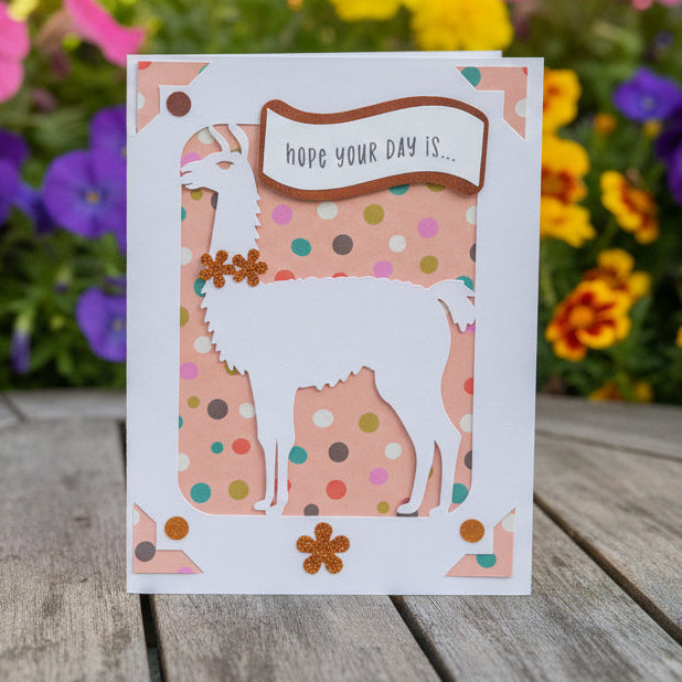 Card with llama design and 'hope your day is' text on a wooden surface with flowers in the background
