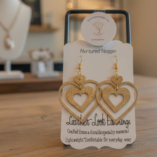 Gold heart-shaped earrings on a display card with 'Nurtured Noggan' branding, placed on a wooden surface.