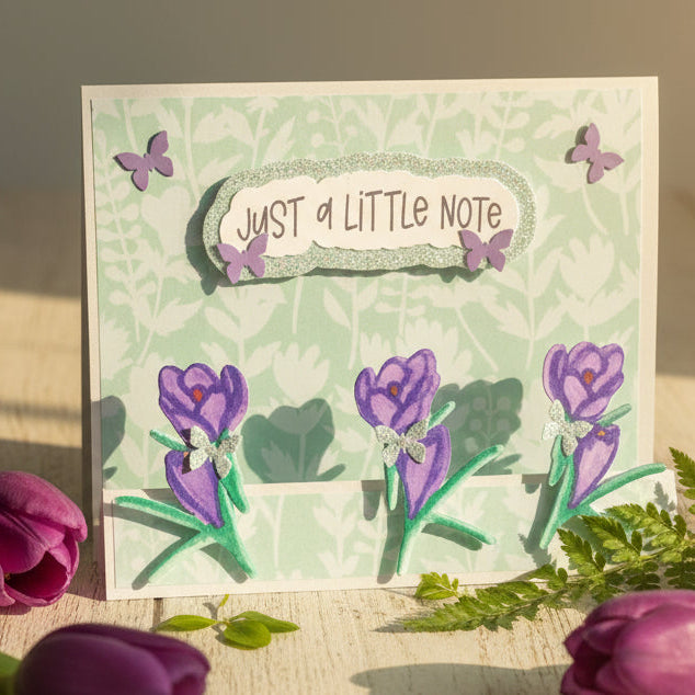 Greeting card with purple flowers and 'Just a Little Note' text on a floral background, surrounded by real purple tulips.