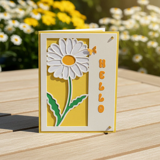 Card with a daisy design and 'hello' text on a wooden surface with flowers in the background