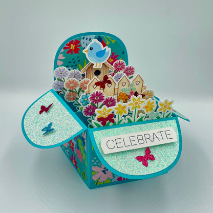 Decorative box with floral design, birdhouse, and 'Celebrate' message on a gray background