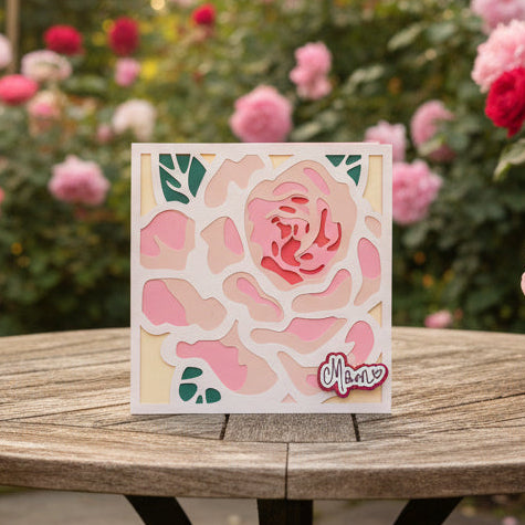 Card with pink rose design on a wooden surface with flowers in the background