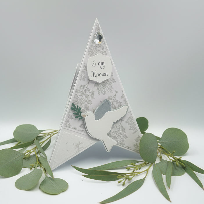Pyramid card with a dove and text, surrounded by greenery on a light gray background