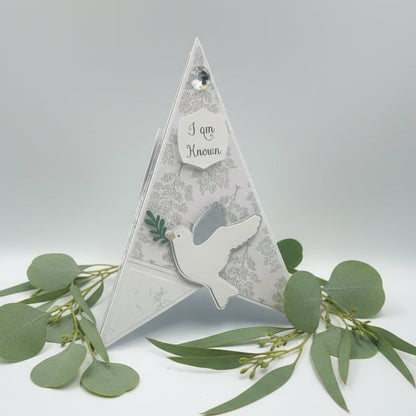 Pyramid card with a dove and text, surrounded by greenery on a light gray background