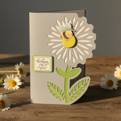 Card with floral design and bee on a wooden surface with daisies