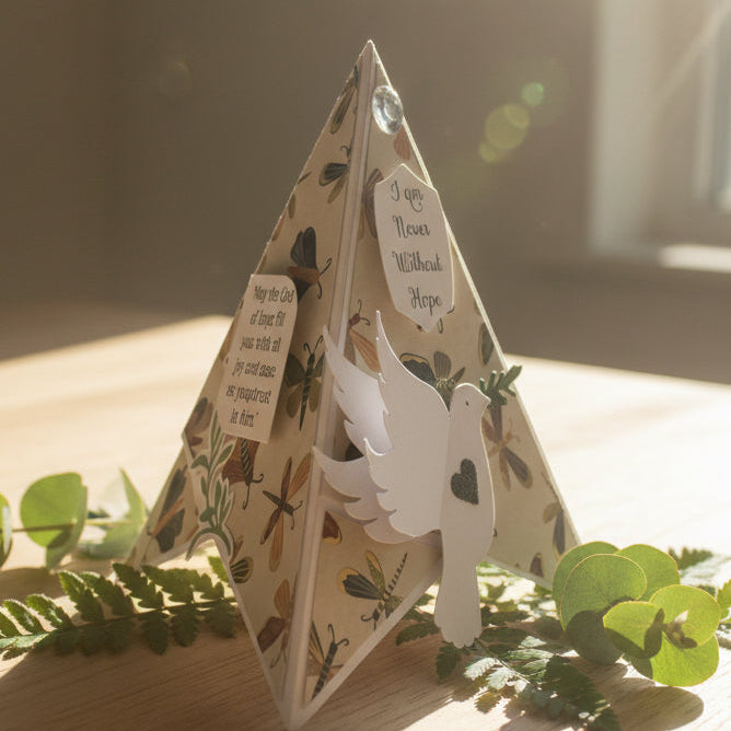 Decorative pyramid card with a dove and text on a wooden surface with greenery