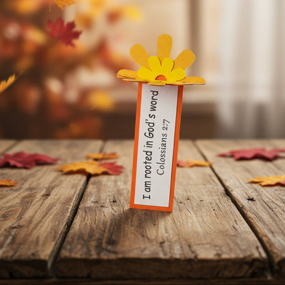 A small decorative Bible bookmark shaped like a yellow flower with orange petals, standing upright on a white card that reads, 'I am rooted in God's word Colossians 2:7' on a light gray background.