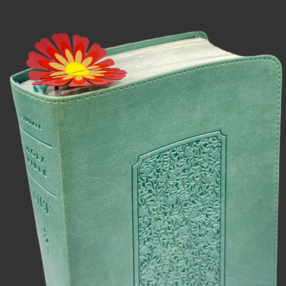 A mint green textured Holy Bible with an embossed floral pattern on the front cover, shown standing upright with a red and yellow daisy flower bookmark tucked behind the top edge.