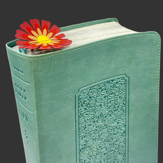 A mint green textured Holy Bible with an embossed floral pattern on the front cover, shown standing upright with a red and yellow daisy flower bookmark tucked behind the top edge.